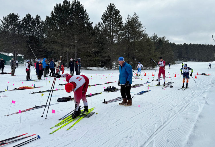 Eric Parrot (14) and Denny Paull (17) are switching skis while Milan Baic (74) and Drew Moore (8) arrive in the Transition Zone at the 2025 Winter Wolf Skiathlon at Forbush Corner..